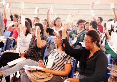 grupo de pessoas está com as mãos levantada, cada qual segurando um papel, durante votação na 1ª Conferência Municipal sobre os Direitos das Pessoas com Deficiência de Paulínia, realizada em 2011 (Tô Dentro!)